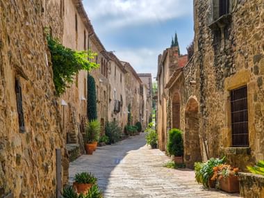 Schmale Kopfsteinpflastergasse in Katalonien, gesäumt von historischen Steinhäusern, Topfpflanzen und einem Kirchturm im Hintergrund.