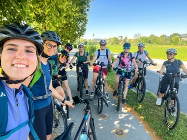 Gruppe von Radfahrern mit Helmen posiert auf einem asphaltierten Weg in Salzburg. Grüne Felder und Bäume im Hintergrund unter blauem Himmel.