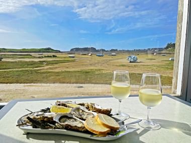 Teller mit frischen Austern, Zitrone und Brot neben zwei Gläsern Weißwein auf einem Tisch mit Blick auf eine Gezichtenbucht mit Booten in der Bretagne.
