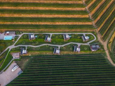 Aerial view of glamping huts arranged along a curved path between agricultural fields near Cividale del Friuli, with a pool visible.