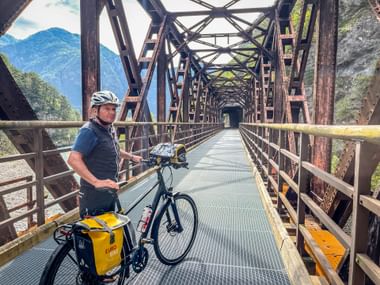 Radfahrer mit Helm und Tourenrad auf historischer Eisenbahnbrücke mit Stahlfachwerk. Berge durch Brückenöffnung bei Chiusaforte sichtbar.
