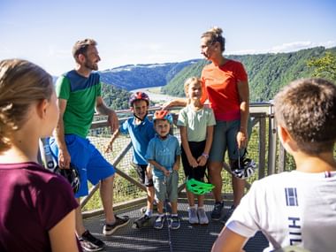 Family with children in cycling helmets on viewing platform at Schlögener Schlinge, overlooking the Danube river bend and green hills.