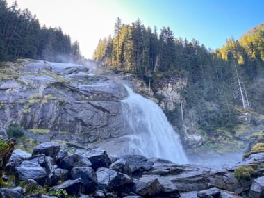 Krimml Waterfalls cascading over rocky cliffs surrounded by forest. Large boulders in foreground, mist rising from the falls.
