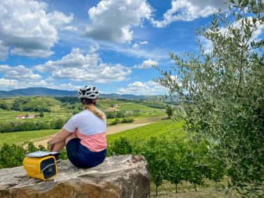 Radfahrerin sitzt auf Steinbank unter Olivenbaum am Rastplatz Ruttar in Collio, Friaul, mit Blick auf grüne Weinberge und Hügel.