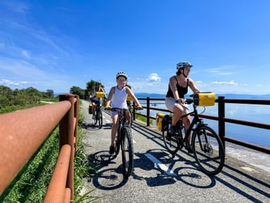Family cyclists with yellow panniers riding on paved path beside Grado Lagoon, with blue water and mountains visible in background.