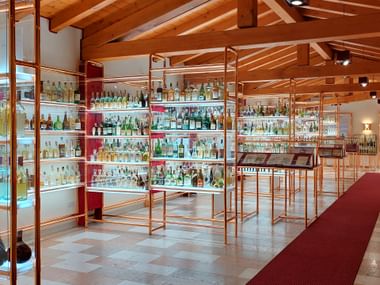 Innenraum des Grappa-Museums mit kupferfarbenen Glasregalen voller Flaschen. Sichtbare Holzbalken an der Decke, roter Teppich am Boden.