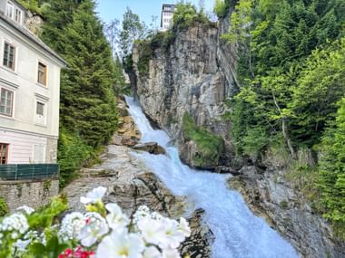 Gasteiner Waterfall cascading through rocky gorge in Bad Gastein, with white building on left and white flowers in foreground.