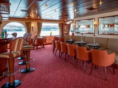 Interior of MS Switzerland bar with orange chairs, round tables, red carpet, and large windows overlooking the water. Wooden ceiling with lights.
