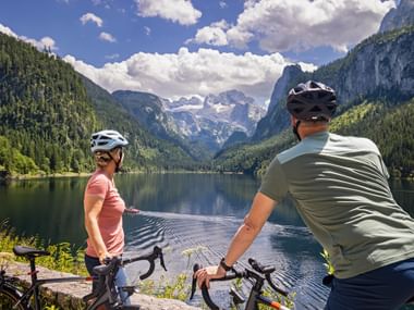 Two cyclists with helmets pause at Gosausee lake shore, viewing the Dachstein mountain range with snow-capped peaks and forested slopes.