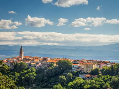 Panoramic view of Vrbnik, a hilltop town on Krk Island, Croatia, with red-tiled roofs and a church tower, surrounded by green trees and mountains.
