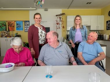 Two female staff members standing behind three seated seniors at a table in a bright community room with yellow walls and colorful artwork.