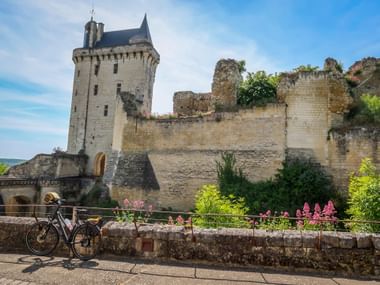 Touring bicycle parked on stone wall with pink flowers in front of Chinon Castle ruins. Medieval tower and crumbling walls under blue sky.