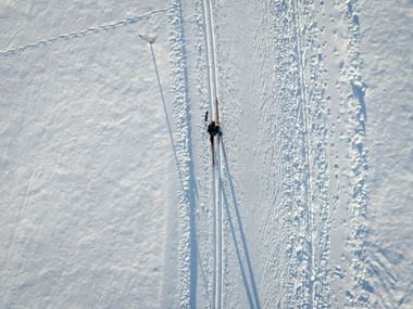 Luftaufnahme eines einzelnen Langläufers auf einer präparierten Schneepiste mit parallelen Spuren und sichtbaren Fußabdrücken in der weißen Landschaft.
