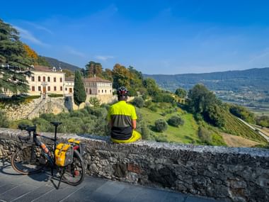 Cyclist in yellow jersey sitting on stone wall overlooking Italian countryside. Touring bike with yellow panniers stands nearby. Historic buildings and hills visible.