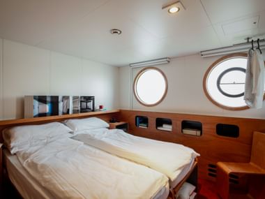 Ship cabin on MS Ave Maria with double bed, white bedding, wooden paneling, two round portholes, and built-in storage compartments.