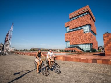 Zwei Radfahrer auf einem Platz vor dem roten Turm des Museum aan de Stroom in Antwerpen unter blauem Himmel.