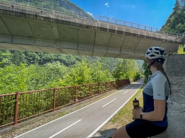 Female cyclist with helmet sitting on cycle path under concrete bridge, holding water bottle. Green vegetation and mountains visible in background.