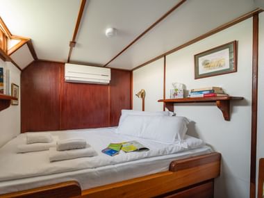 Kompakte Schiffskabine auf der MS Caprice mit Doppelbett, weißer Bettwäsche, Holzverkleidung, Klimaanlage, Regal mit Büchern und kleinem Fenster.