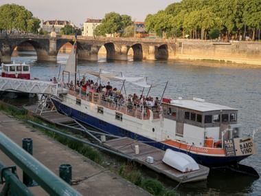 Passenger boat with bar docked on Loire River in Angers. Historic stone bridge with arches visible in background, tree-lined promenade on right.