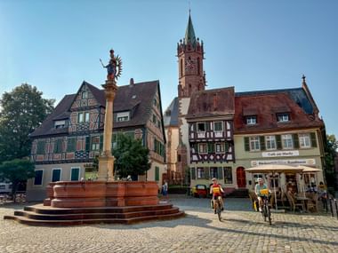 Ladenburger Marktplatz mit verziertem Brunnen, Fachwerkhäusern und Kirchturm. Radfahrer in gelben Westen fahren über das Kopfsteinpflaster.