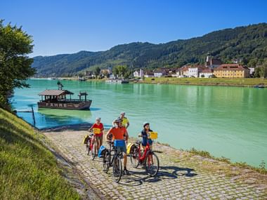 Four cyclists with bikes at riverside waiting for ferry boat on turquoise Danube River. Colorful buildings and forested hills in background.