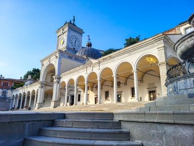 Weißes neoklassizistisches Gebäude mit Arkadengang und Uhrturm in Udine, Italien. Steintreppen führen zur beleuchteten Arkade unter blauem Himmel.