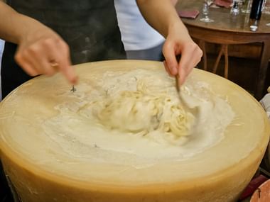 Hands mixing fresh pasta inside a hollowed Tuscan cheese wheel. The creamy interior shows pasta being stirred with steam rising from the dish.