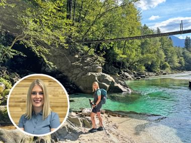 Female hiker with backpack standing on rocky shore beside turquoise Soča River. Suspension bridge and forested mountains visible in background.