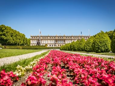 Schloss Herrenchiemsee in Bayern mit leuchtend rosa Blumenbeeten im Vordergrund, eingerahmt von gepflegten Rasenflächen und Bäumen.