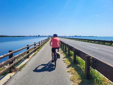 Radfahrer im rosa Shirt auf Radweg entlang Damm zwischen blauer Lagune und Straße bei Grado auf dem Alpe-Adria-Radweg.