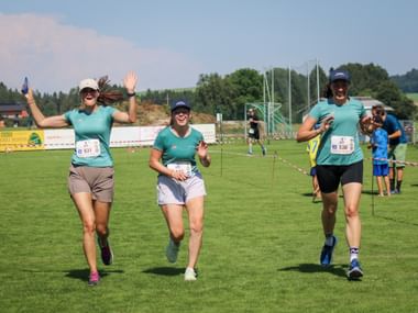 Three female runners in turquoise shirts crossing finish line at Grabenseelauf 2026, waving and celebrating on grass field with spectators.