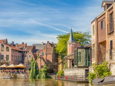 Historic brick buildings along the Leie River in Ghent with a church spire, modern glass structure, and waterfront terrace under blue sky.