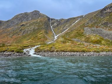 Zwei Wasserfälle stürzen von felsigen Bergen in den Bygdin-See im Jotunheimen, Norwegen. Grüne und braune Hänge treffen auf türkisfarbenes Wasser.