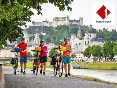 Stadt Salzburg mit 3 Radfahrern an der Salzach und der Festung im Hintergrund