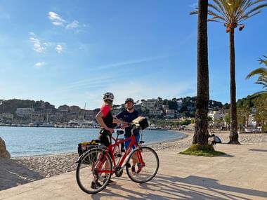Zwei Radfahrer mit roten Rädern machen Pause an der Promenade am Strand von Port de Sóller auf Mallorca, mit Palmen, Hafen und Hanghäusern.