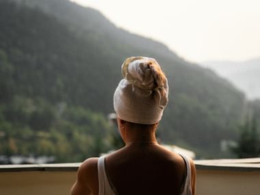 Rear view of person with white towel wrapped on head, looking at misty mountain landscape from balcony after sauna.