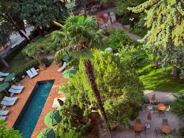 Aerial view of a lush hotel garden in Bolzano with a rectangular pool, white sun loungers, palm trees, and a terrace with tables and umbrellas.