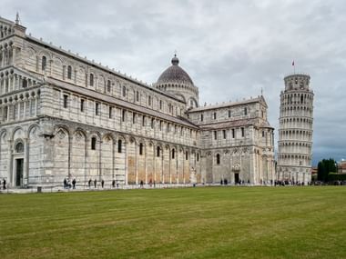 Piazza dei Miracoli in Pisa showing the white marble Cathedral with its dome, the famous Leaning Tower, and green lawn under cloudy sky.