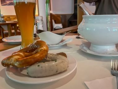 Two white sausages with pretzel on a plate, served with wheat beer and white sauce in a tureen. Traditional Bavarian breakfast setting.