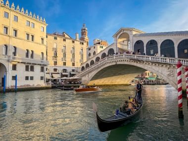 Gondola with passengers on Canal Grande in Venice, with the historic Rialto Bridge and traditional buildings under blue sky.