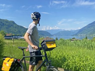 Radfahrer mit Helm steht neben Fahrrad mit gelben Taschen, Blick auf Ossiacher See mit Bergen im Hintergrund unter blauem Himmel.