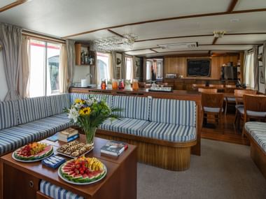 Innenraum der MS Caprice mit blau gestreiften Sitzgelegenheiten, Holzcouchtisch mit Obsttellern, Blumen und offener Küche mit Holzverkleidung.