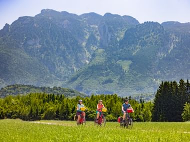 Three cyclists riding through a green meadow near Salzburg-Wals with the forested Untersberg mountain range in the background under clear skies.