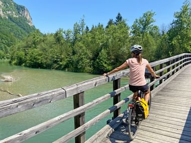 Radlerin in rosa Shirt mit Helm auf Holzbrücke über türkisfarbener Drau in Kärnten, umgeben von grünem Wald und Bergen.