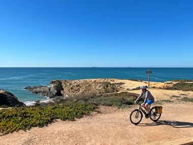 Cyclist riding along sandy coastal path in Porto Covo, Portugal. Rocky cliffs and turquoise ocean visible under clear blue sky.