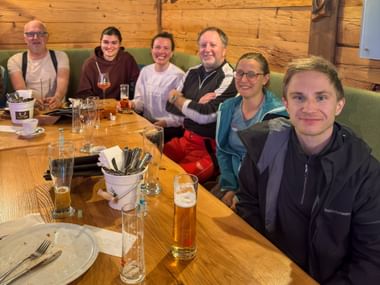Sechs Personen sitzen an einem Holztisch in einer Berghütte mit Biergläsern und leeren Tellern. Holzvertäfelte Wände und grüne Sitzbank sichtbar.