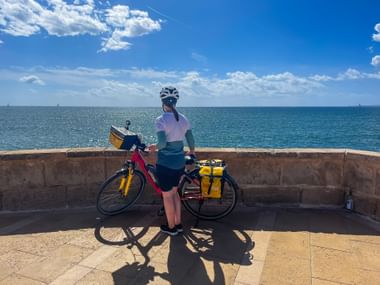 Radfahrerin mit Helm steht neben Tourenrad mit gelben Packtaschen auf Steinterrasse mit Blick auf das türkisfarbene Mittelmeer bei Sóller.