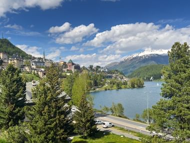 Malerischer Blick auf St. Moritz mit blauem See, umliegenden Stadtgebäuden, grünen Wäldern und schneebedeckten Alpen unter blauem Himmel mit Wolken.