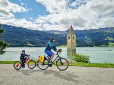 Erwachsener und Kind radeln am Uferweg des Reschensees mit dem berühmten versunkenen Kirchturm im türkisfarbenen Wasser, Berge im Hintergrund.