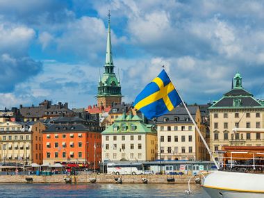 Blick auf die alten Häuser und den Kirchturm von Strandvägen in Stockholm mit der schwedischen Fahne im Vordergrund